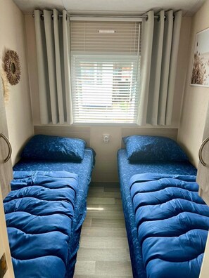 Une chambre de taille modeste, équipée de deux lits, d’une fenêtre avec stores et d’un petit luminaire fixé au mur.