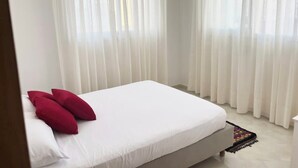 A neatly made bed with white sheets and two red pillows, a white headboard, and sheer white curtains. - Apartment 5 minutes' walk from the beach (martil)