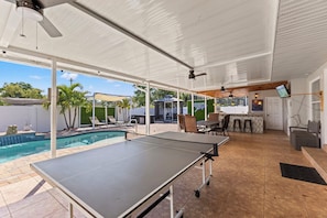 An outdoor area with a ping pong table, a pool, and a covered seating area with chairs and tables.