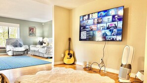 A living room with a large flat-screen TV mounted on the wall displaying various shows, a guitar leaning against the wall, a fluffy white rug on the floor, and a small speaker on the right. - Cozy, Quiet, Bothell River House (Bothell)