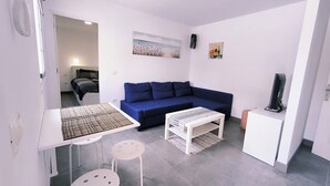 Un espacio habitable compacto con un sofá azul, una mesita blanca, dos sillas blancas y un televisor sobre un mueble.