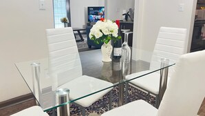 Ein Essbereich mit Glastisch und weißen Stühlen, einer Vase mit weißen Blumen und einer Weinflasche.