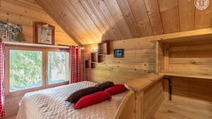 Ein Zimmer einer Holzhütte mit einem Bett, roten Kissen, einem Fenster mit Vorhängen und einer kleinen Ablage mit einem Bild.