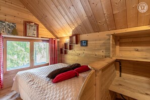 Une chambre dans un chalet en bois, avec un lit, des oreillers rouges, une fenêtre avec des rideaux et une petite étagère sur laquelle est posée une photo.