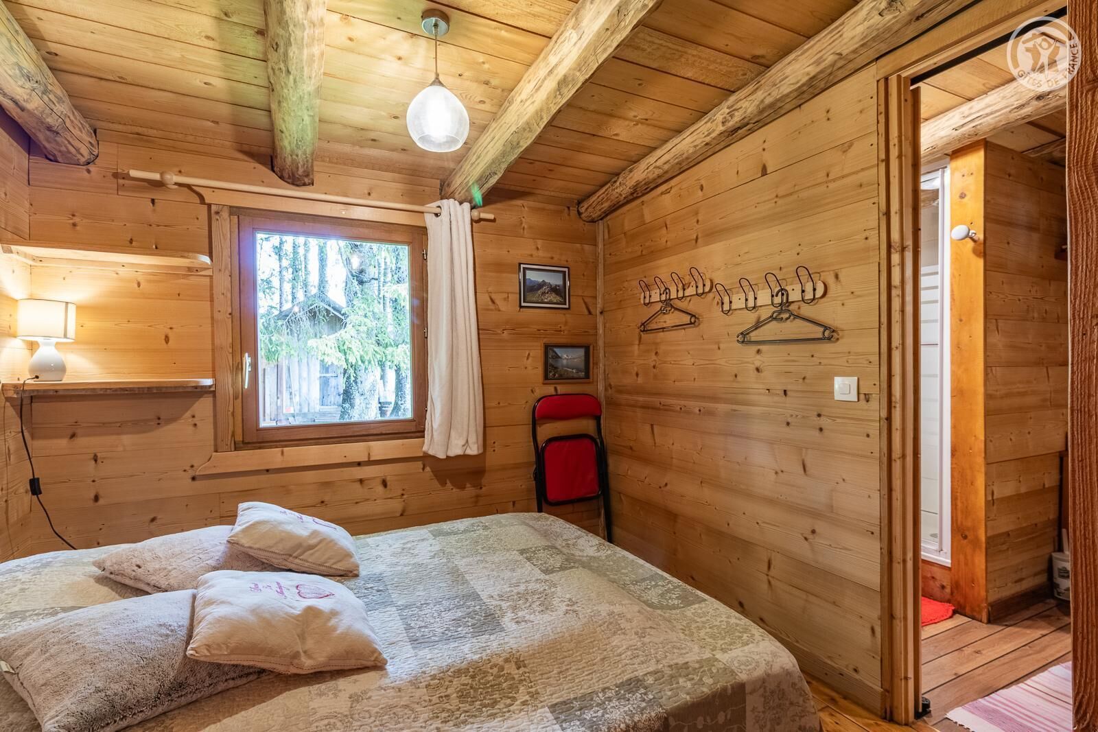 A wooden cabin room with a bed, a window, a hanging light fixture, and a coat rack.