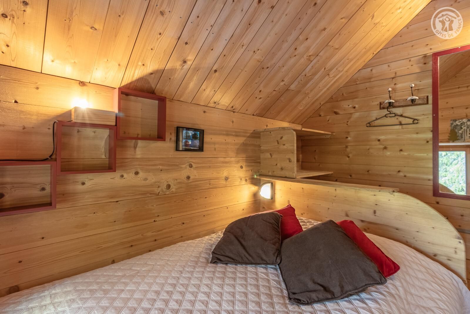 Een slaapkamer in een houten chalet met een bed, kussens, een hanglamp en een klein raam.
