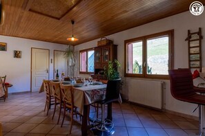 A dining area with a wooden table set for four, surrounded by chairs. There is a radiator under a window, and a plant is visible in the background.