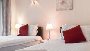 Habitación de hotel con dos camas, cada una con ropa blanca y almohadas rojas, una mesita de noche con lámpara, y una lámpara de pared.