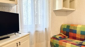A room with a television on a white cabinet, a colorful sofa, a window with sheer curtains, and a white shelf above the sofa. - RM11 - Senigallia, three-room apartment facing the sea (Senigallia)