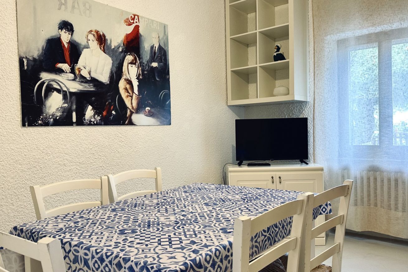 A dining area with a table covered in a patterned tablecloth, a television on a cabinet, and a painting on the wall.