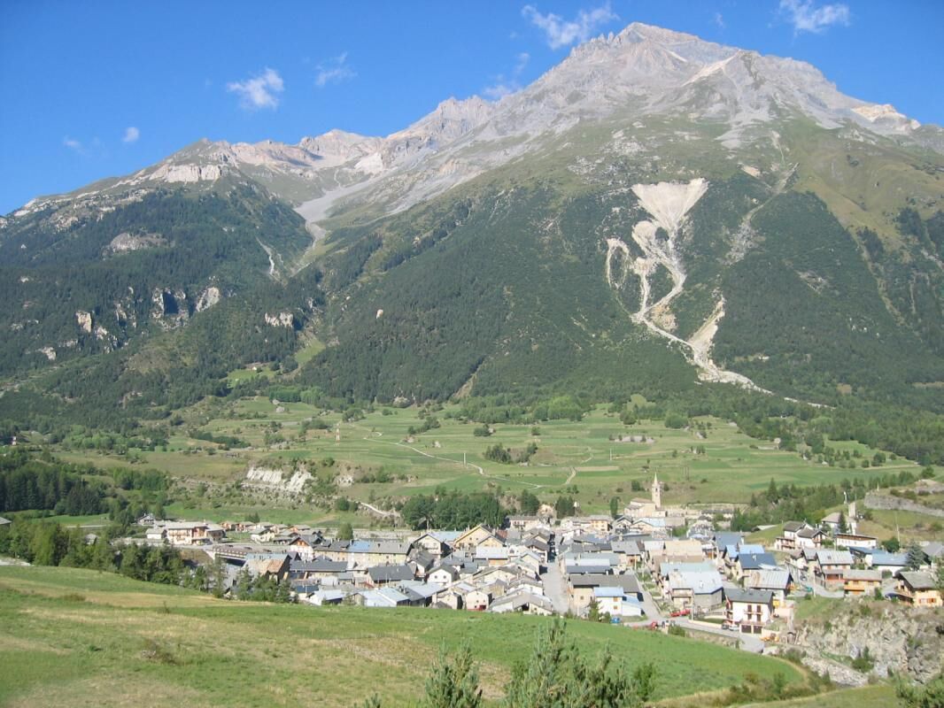 Un pueblo de montaña con edificios agrupados al pie, rodeado de colinas verdes y una montaña destacada en el fondo.