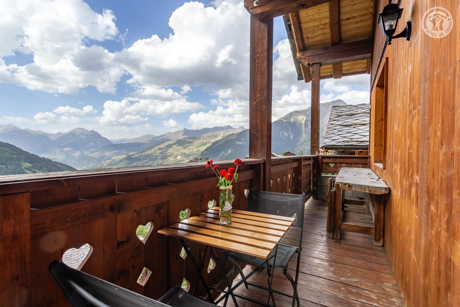 Eine Holzterrasse mit Tisch und Stühlen, einem Blumenvasen und Blick auf die Berge.