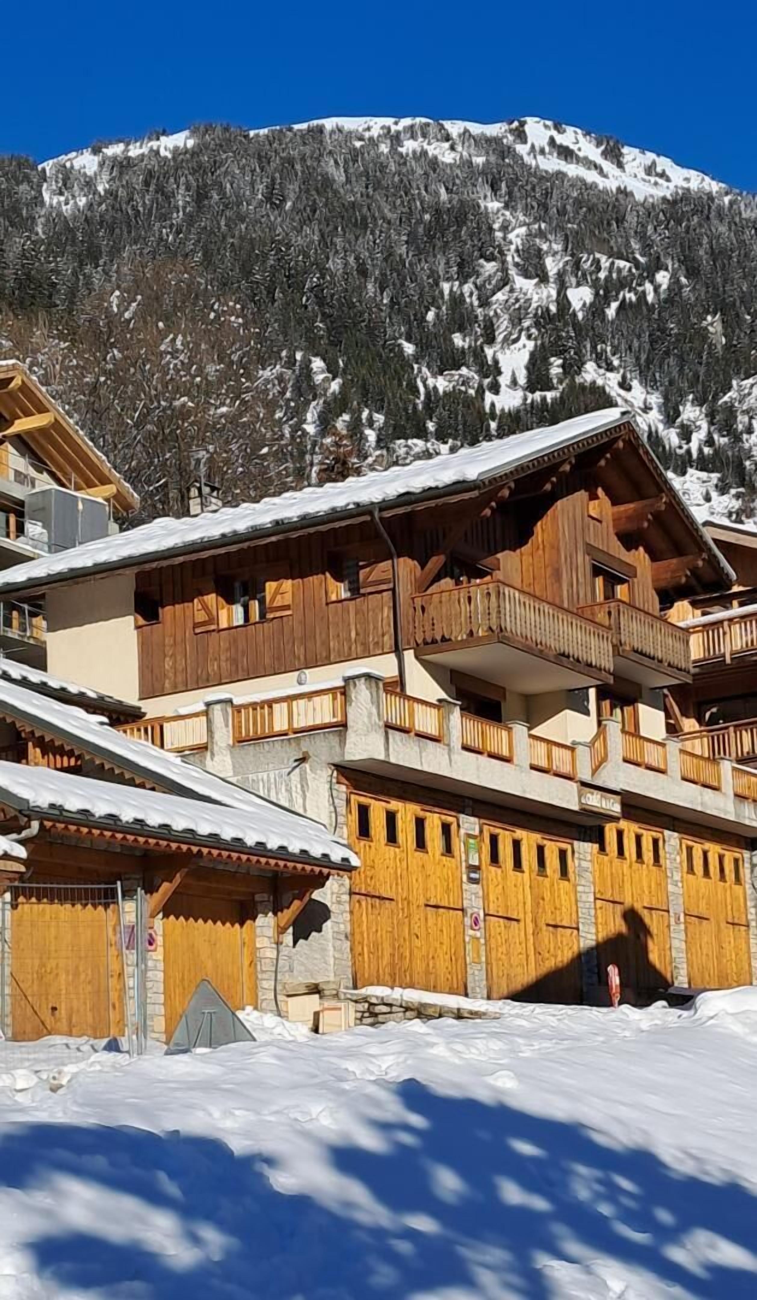 A multi-story wooden chalet with balconies, set against a snowy mountain backdrop.