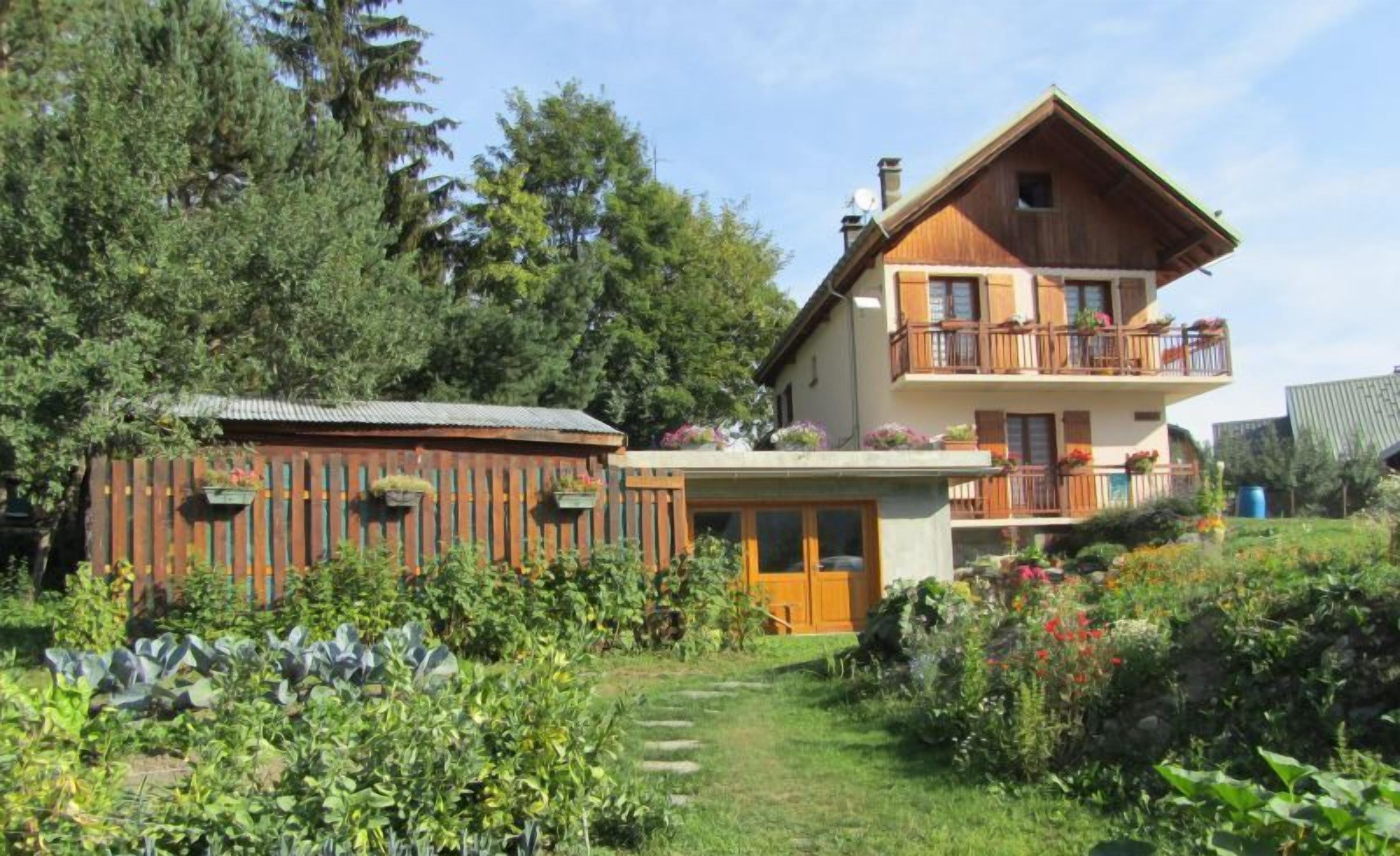 A two-story wooden house with a balcony, surrounded by lush greenery and a garden.