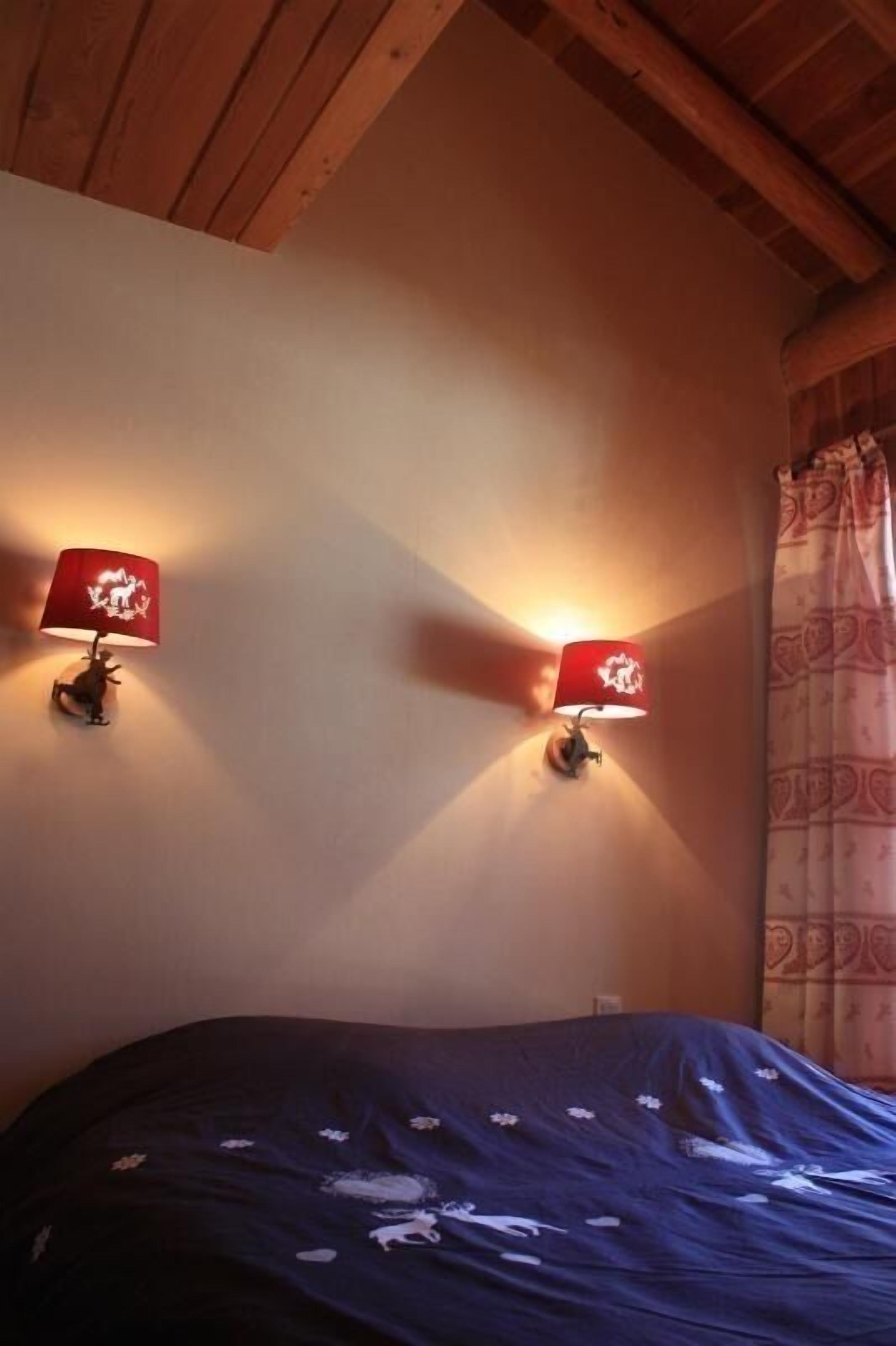 Una camera da letto con soffitto in legno, due lampade a parete con paralumi rossi, un letto con copripiumino blu scuro e una tenda a fantasia.