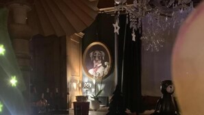 A cozy room with a wooden ceiling, a chandelier, a framed portrait, and a teddy bear on a table.
