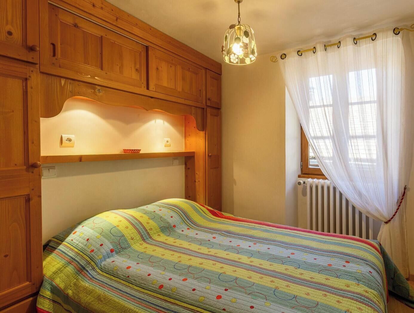 A bedroom with a wooden headboard, a bed with a colorful striped bedspread, a hanging light fixture, and a window with sheer curtains.