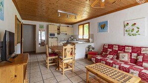 Una sala de estar acogedora con techo de madera, un televisor, una mesa de madera, un sofá con estampado rojo y blanco y una cocina al fondo.