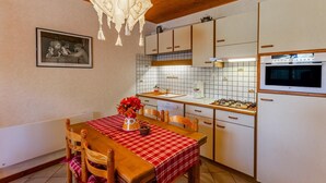 A kitchen with wooden cabinets, a tiled backsplash, a dining table with a red checkered tablecloth, and a chandelier. - LE TALAPET - CONFETTI (ALBIEZ MONTROND)