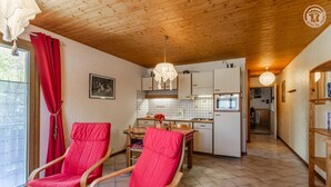 A room with two red chairs, a wooden ceiling, a kitchen area with white cabinets, and a dining table with a red tablecloth. - LE TALAPET - CONFETTI (ALBIEZ MONTROND)