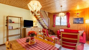 A cozy living room with a wooden ceiling, a staircase, a red sofa, a dining table with a checkered tablecloth, and a television on a wooden cabinet. - LE TALAPET - CONFETTI (ALBIEZ MONTROND)