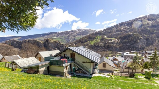 Ein Bergdorf mit traditionellen Häusern, ein markantes Haus mit Balkon und ein Tal mit Bergen im Hintergrund.