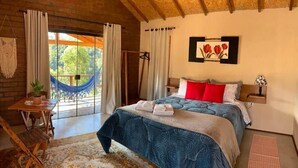 Une chambre à coucher avec un lit, une table de chevet, une plante suspendue, une fenêtre avec des rideaux et un hamac à l’extérieur.