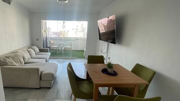 Una sala de estar moderna con un sofá, una mesa de comedor con sillas, un televisor montado en la pared y vistas al balcón exterior.
