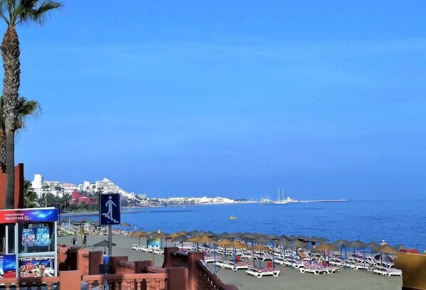 A beach with rows of sun loungers and umbrellas, a clear blue sky, and a distant view of a cityscape.
