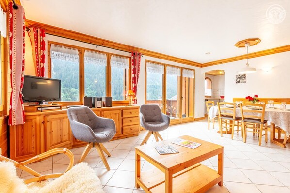 A cozy room with a TV, two grey chairs, a wooden table, and a dining area with chairs and a table.