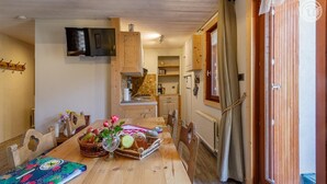 A wooden dining table with a floral centerpiece, a TV mounted on the wall, and a kitchen area with wooden cabinets and a window.