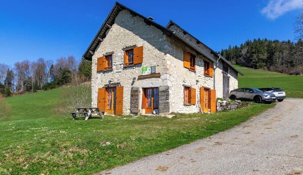 Un bâtiment en pierre avec des volets en bois, une table de pique-nique et des voitures garées.