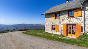 Una casa de piedra con contraventanas de madera, un camino de grava y vistas a colinas distantes.