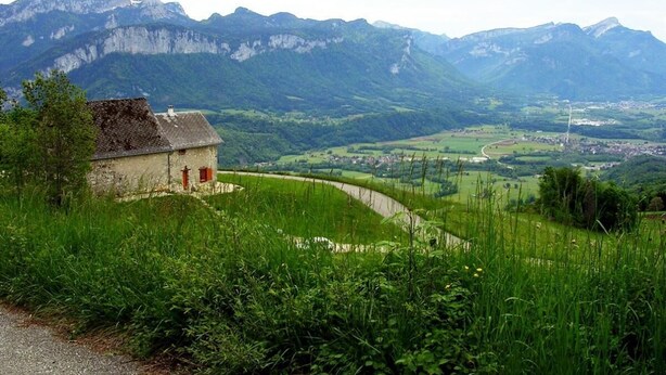 Un paisaje rural con una casa en una ladera, un camino sinuoso y montañas al fondo.