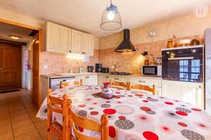 Een keuken met houten kastjes, een eettafel met een tafelkleed met patroon, een hanglamp en een magnetron.