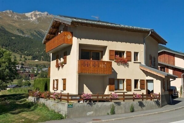 A two-story building with wooden balconies and flower boxes, set against a mountainous backdrop.
