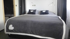 A bedroom with a bed, two pillows with heart designs, a wall-mounted light fixture, and a shower area visible through an open door. - LE REFUGE DU BACHAL N°1 (NOTRE DAME DE BELLECOMBE)