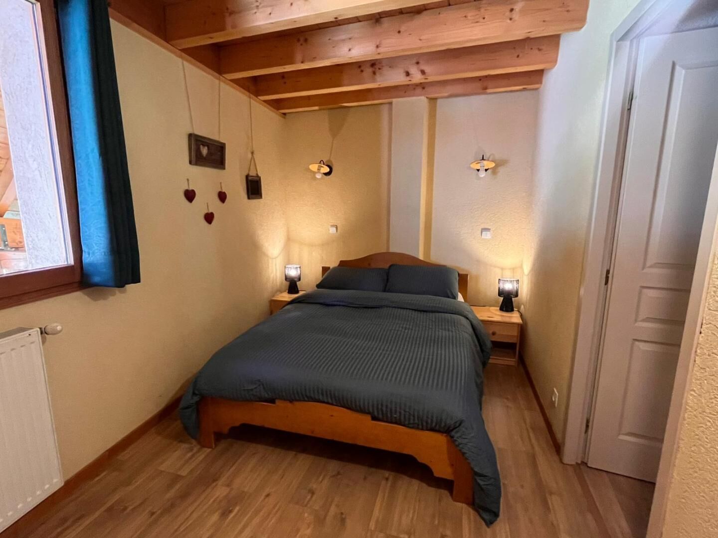 A bedroom with a wooden bed, two bedside lamps, a window with blue curtains, and a wall with heart decorations.