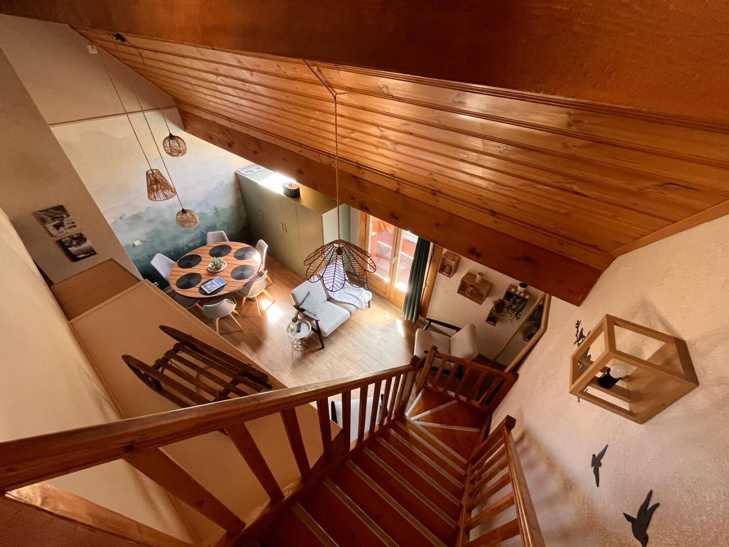 Una escalera de madera conduce a un área de comedor con una mesa redonda y sillas, un sofá y una lámpara colgante.