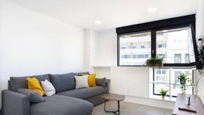 Un salon moderne avec un canapé d’angle gris, une petite table basse et une fenêtre donnant sur des immeubles.