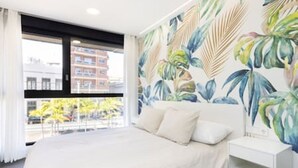 Une chambre avec un grand lit, une table de chevet et une fenêtre donnant sur des immeubles et des arbres.