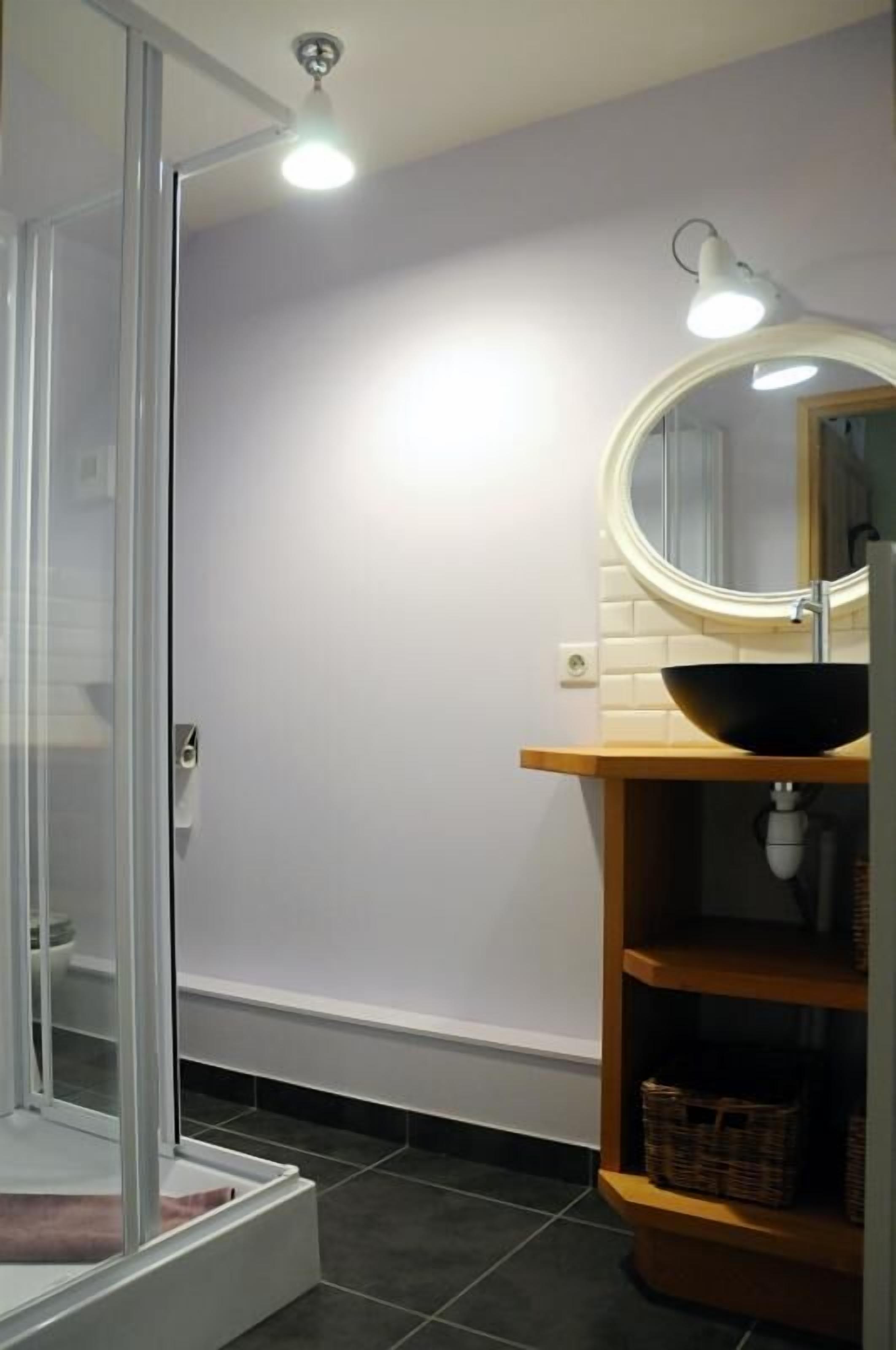A bathroom with a glass shower enclosure, a round mirror, a black sink, and a wooden shelf with baskets.