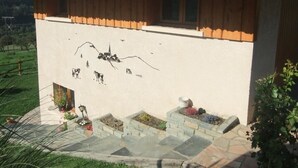 Ein Gebäude mit weißer Fassade und einem Holzteile im oberen Bereich. Auf der Wand befindet sich ein Wandbild, das eine Berglandschaft, eine Kirche und Kühe zeigt.