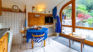 A cozy kitchen with a dining table, wooden cabinets, a clock, and a view of a garden through a large window. - L'ARCHE - RUFFIEUX - Gîte (RUFFIEUX)