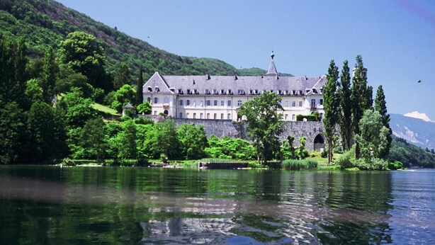 A large, historic building with multiple windows and a prominent tower, surrounded by lush greenery and a calm lake. - L'ARCHE - RUFFIEUX - Gîte (RUFFIEUX)
