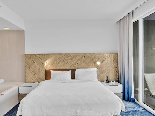 A modern hotel room with a large bed, wooden headboard, bedside tables, and a view through a glass door.