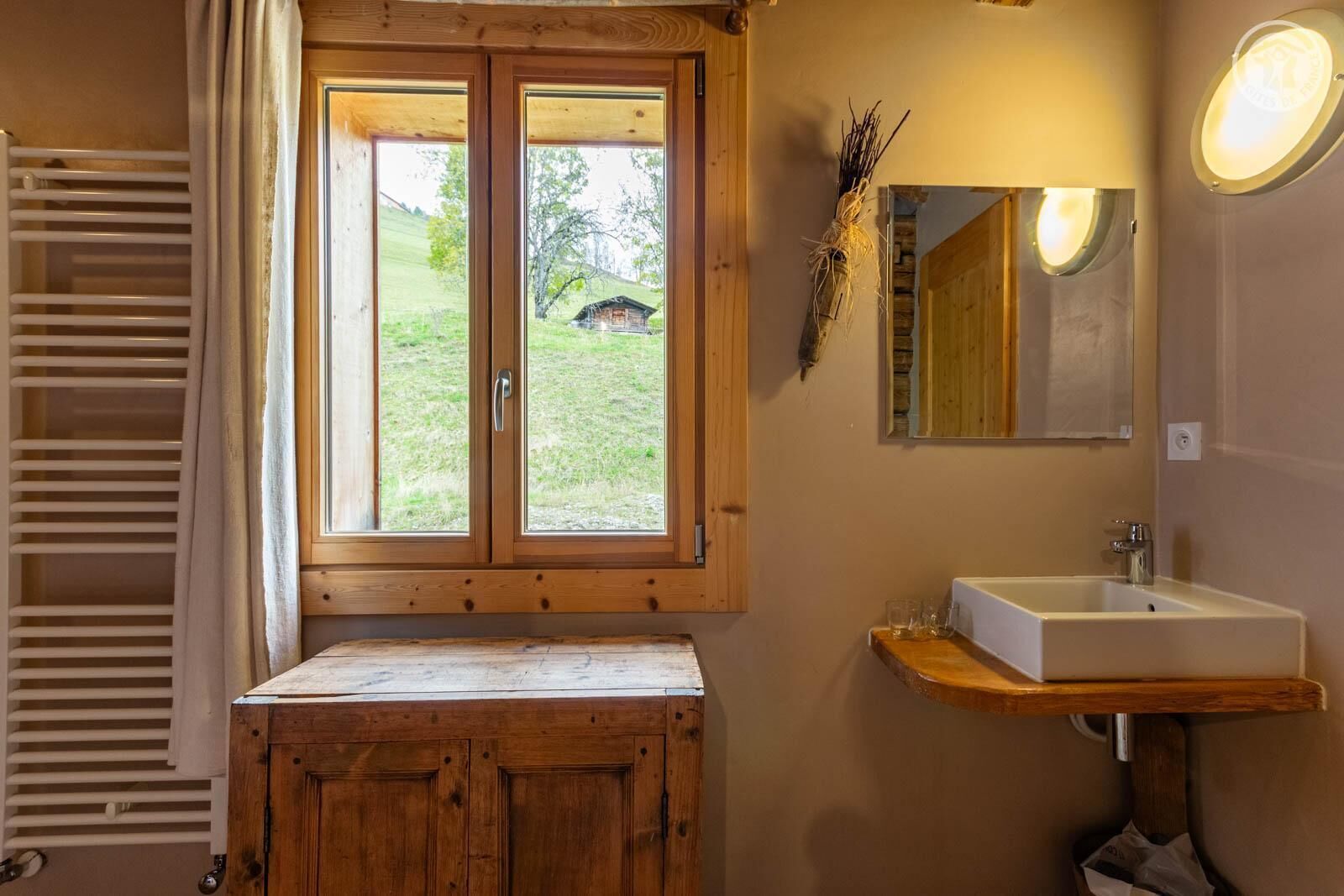 A bathroom with a wooden vanity, a sink, a mirror, and a window with a view of a house and greenery.