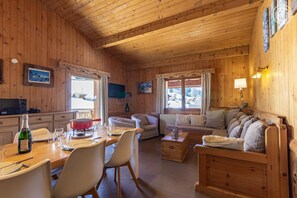 A wooden interior with a dining table, chairs, a sofa, a television, and a window with curtains.