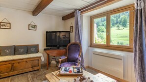 A cozy room with a wooden ceiling, a sofa with heart-patterned cushions, a purple armchair, a television, a table with a teapot, and a window with a view of greenery. - LA COMBE DE MOULIN - PEISEY NANCROIX (PEISEY NANCROIX)