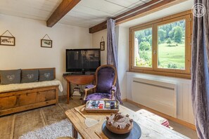 Een gezellige kamer met een houten plafond, een bank met kussens met hartjesprint, een paarse fauteuil, een televisie, een tafel met een theepot en een raam met uitzicht op groen.